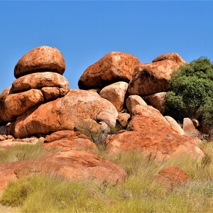 May include: A collection of large, red, rounded boulders in a desert landscape. The boulders are stacked on top of each other, and there is a small green bush growing on one of the boulders.