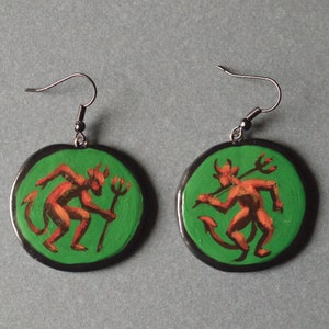 Peut inclure: Une paire de boucles d'oreilles avec des pendentifs circulaires verts représentant des figures de diables rouges tenant des tridents. Les boucles d'oreilles ont des crochets argentés.