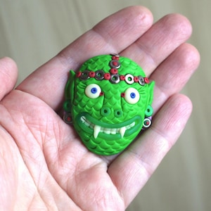 May include: Green and red clay sculpture of a mythical creature with fangs, large eyes, and a crown.