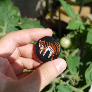 Puede incluir: Una mano sosteniendo un pequeño botón redondo negro con una imagen pintada de un tigre en naranja, negro y blanco.