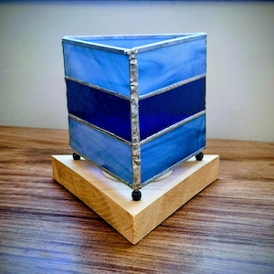 May include: A stained glass candle holder in a triangular shape, featuring shades of blue. The glass is encased in a silver-colored metal frame and sits atop a wooden base with small black feet.