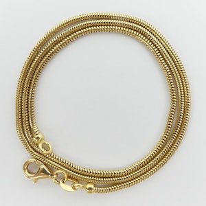 Könnte beinhalten: Eine goldfarbene Schlangenkette mit einem Karabinerverschluss. Die Kette ist kreisförmig gewickelt und zeigt ihr aufwendiges, strukturiertes Design. Die Halskette besteht aus einem glänzenden, metallischen Material.