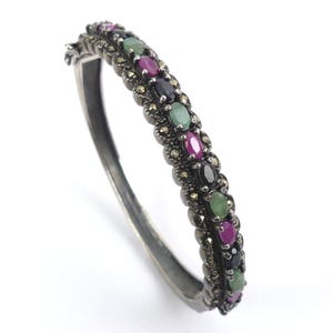 Op de afbeelding: Een scharnierende armband in een donkere metaalafwerking, voorzien van een centrale rij met afwisselend ovaal geslepen groene, roze en zwarte stenen. De band is geaccentueerd met kleine, getextureerde metalen kralen rondom de stenen.