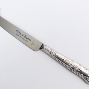 Op de afbeelding: Een roestvrijstalen kaasmes met een decoratief handvat. Het lemmet heeft een gebogen punt met twee tanden. De tekst "Cheese Knife" en "Sheffield England" staat op het lemmet.