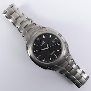 Peut inclure: Une montre Citizen Eco-Drive argentée avec un cadran noir et un affichage de la date. La montre est dotée d'un bracelet en titane. Les aiguilles et les marqueurs de la montre sont argentés.