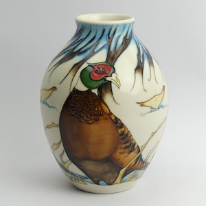 May include: A white ceramic vase with a brown and gold pheasant design. The pheasant is standing in a snowy forest scene with blue and white branches.