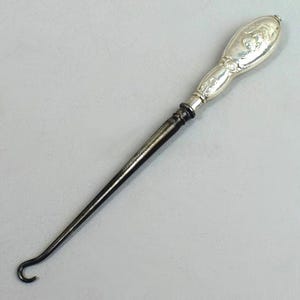 Puede incluir: Un calzador de plata con un mango adornado y un gancho de metal negro. El mango está decorado con un diseño floral.