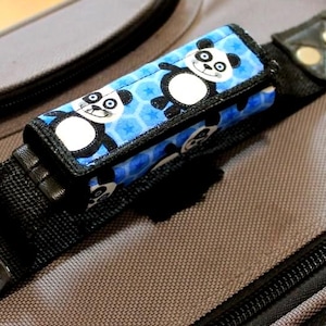 May include: A luggage handle wrap with a blue panda pattern. The wrap is designed to fit around a suitcase handle and features a repeating pattern of panda bears on a blue background. The text "PETITE POIGNEE DE BAGAGE PANDAS" is visible.