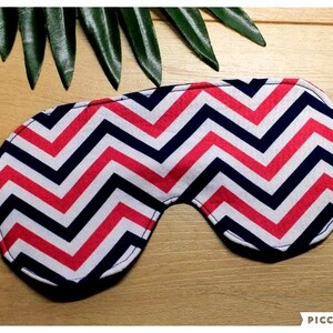 May include: A sleep mask with a chevron pattern in red, white, and navy blue. It features an elastic strap and is designed to cover the eyes. The background includes green leaves on a wooden surface.
