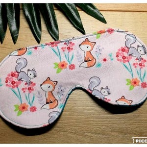 May include: A light pink sleep mask with a repeating pattern of cartoon foxes and squirrels amongst pink flowers and green leaves. The mask has a white elastic band and a teal interior lining. Designed for eye coverage and relaxation.