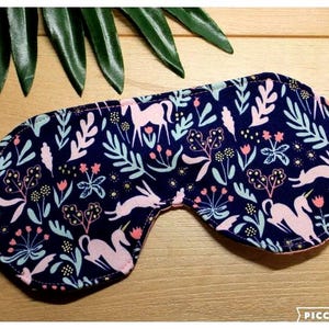 May include: A dark blue sleep mask featuring a repeating pattern of light pink unicorns, rabbits, and various botanical elements in shades of light blue, pink, and yellow. The mask has a contoured shape with an elastic strap on each side.