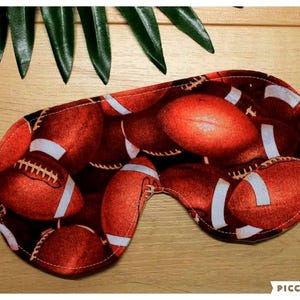 Peut inclure: Un masque de sommeil orné d'un motif répété de ballons de football américains rouges et blancs. Le masque est doté d'un élastique blanc et est conçu pour bloquer la lumière. Fond en bois brun clair.