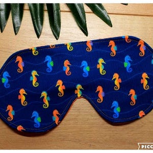 May include: A navy blue sleep mask featuring a pattern of colourful seahorses. The seahorses are in shades of orange, yellow, and blue. The mask has an orange interior and an elasticated band.