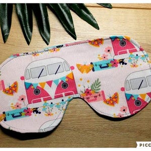 May include: A light pink sleep mask featuring a repeating pattern of a retro camper van, butterflies, and flowers. The mask has a white elastic strap. The camper vans are pink and white.