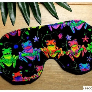 May include: A black sleep mask decorated with a colourful frog pattern. The mask displays frogs in green, blue, pink, and orange, set against a black backdrop. The frogs are positioned on branches with flowers and vines.