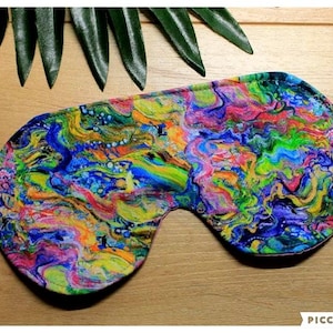 May include: A vibrant sleep mask featuring an abstract design in blue, green, yellow, pink, and purple. It has an elastic strap and is designed to block light. The background is a wooden surface with green leaves.