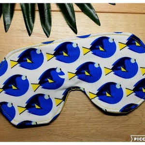 May include: A white sleep mask patterned with blue fish and yellow fins. The mask has a yellow stitched border and an elastic strap. The background is a wooden surface with green leaves.