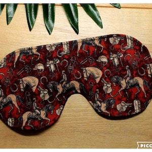 May include: A burgundy sleep mask with a black elastic strap. The mask displays a pattern of horses, riders, and equestrian equipment in cream and black. It rests on a wooden surface with green leaves.