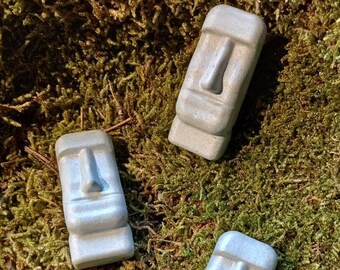Könnte beinhalten: Drei kleine graue Betonstatuen von Osterinselköpfen, jede mit einem eigenen Gesichtsausdruck, sind auf einem Bett aus grünem Moos angeordnet.