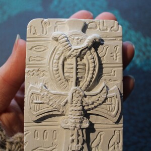 K&ouml;nnte beinhalten: Wei&szlig;e Gipsreliefskulptur eines &auml;gyptischen Ankh-Symbols mit einer Kobra und Fl&uuml;geln. Das Ankh ist von Hieroglyphen umgeben.