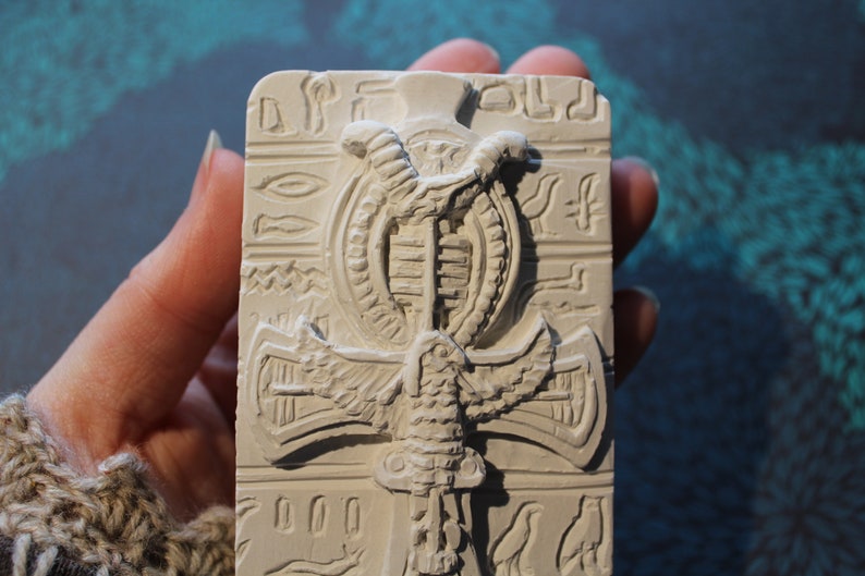 K&ouml;nnte beinhalten: Wei&szlig;e Gipsreliefskulptur eines &auml;gyptischen Ankh-Symbols mit einer Kobra und Fl&uuml;geln. Das Ankh ist von Hieroglyphen umgeben.