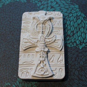 K&ouml;nnte beinhalten: Wei&szlig;er Ankh-Anh&auml;nger aus Keramik mit einem gefl&uuml;gelten Skarab&auml;us-Design. Der Anh&auml;nger ist von Hieroglyphen umgeben.