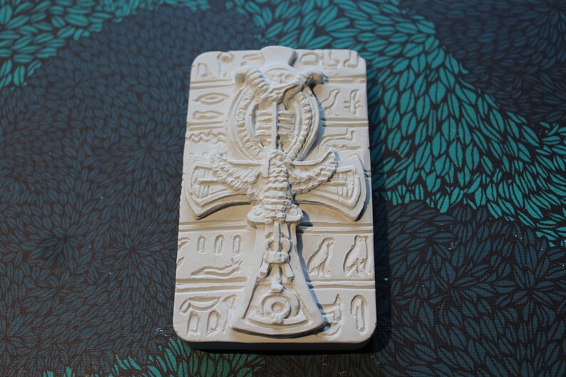 K&ouml;nnte beinhalten: Wei&szlig;er Ankh-Anh&auml;nger aus Keramik mit einem gefl&uuml;gelten Skarab&auml;us-Design. Der Anh&auml;nger ist von Hieroglyphen umgeben.