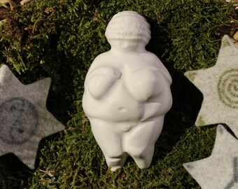 Könnte beinhalten: Weiße Keramikfigur einer Frau mit rundem Körper und einem einfachen, stilisierten Design. Die Figur steht auf einem Bett aus grünem Moos.