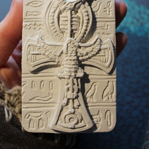 K&ouml;nnte beinhalten: Wei&szlig;er Ankh-Anh&auml;nger aus Keramik mit einem gefl&uuml;gelten Skarab&auml;us-Design. Der Anh&auml;nger ist mit Hieroglyphen und anderen &auml;gyptischen Symbolen verziert.