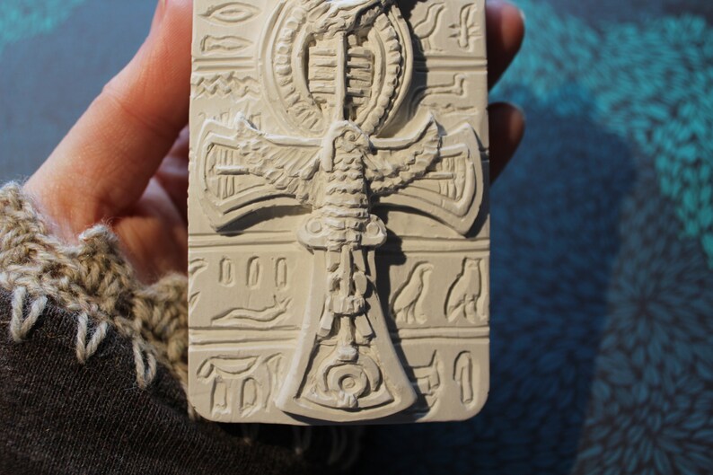 K&ouml;nnte beinhalten: Wei&szlig;er Ankh-Anh&auml;nger aus Keramik mit einem gefl&uuml;gelten Skarab&auml;us-Design. Der Anh&auml;nger ist mit Hieroglyphen und anderen &auml;gyptischen Symbolen verziert.