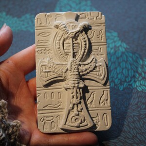 K&ouml;nnte beinhalten: Ein wei&szlig;er Ankh-Anh&auml;nger aus Keramik mit aufwendigen Details, darunter eine gefl&uuml;gelte Schlange und Hieroglyphen. Der Anh&auml;nger wird in einer Hand gehalten.