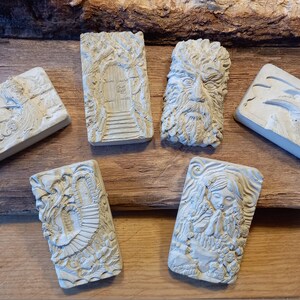 K&ouml;nnte beinhalten: Sechs kleine, rechteckige, wei&szlig;e Betonskulpturen mit verschiedenen Designs. Die Skulpturen zeigen einen Baum mit einer T&uuml;r, ein Gesicht mit Bart, einen Vogel mit Schnabel, ein Spiralmuster und eine Frau mit gefalteten H&auml;nden zum Gebet.