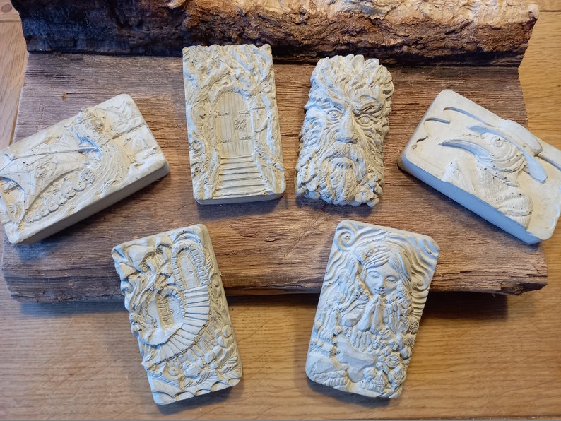 K&ouml;nnte beinhalten: Sechs kleine, rechteckige, wei&szlig;e Betonskulpturen mit verschiedenen Designs. Die Skulpturen zeigen einen Baum mit einer T&uuml;r, ein Gesicht mit Bart, einen Vogel mit Schnabel, ein Spiralmuster und eine Frau mit gefalteten H&auml;nden zum Gebet.