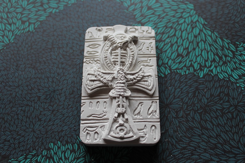 K&ouml;nnte beinhalten: Wei&szlig;er Keramik-Ankh-Anh&auml;nger mit Hieroglyphen und einem gefl&uuml;gelten Skarab&auml;us-Design.