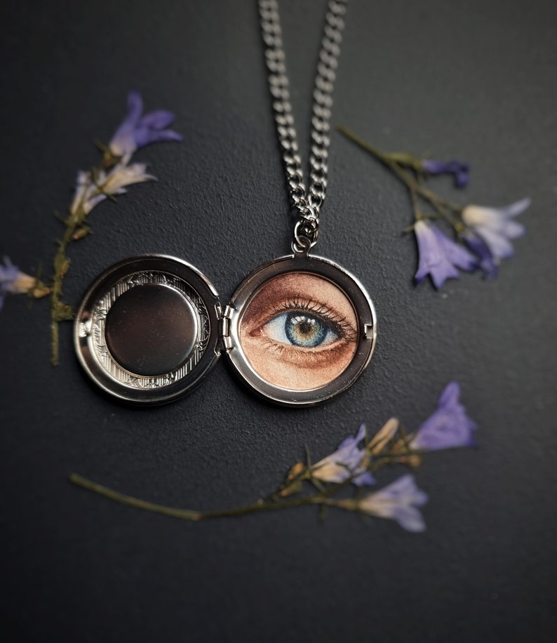 Op de afbeelding: Zilveren ketting met een medaillon hanger met een gedetailleerde schildering van een blauw en bruin oog. Het medaillon is open en toont het kunstwerk. Gedroogde paarse bloemen zijn rond de ketting op een donkere achtergrond geplaatst.