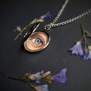 Op de afbeelding: Een zilveren ketting met een ovale hanger met een gedetailleerde schildering van een blauw en bruin oog. Het medaillon is open en onthult het kunstwerk. De ketting ligt op een donkere ondergrond, met takjes paarse bloemen verspreid.