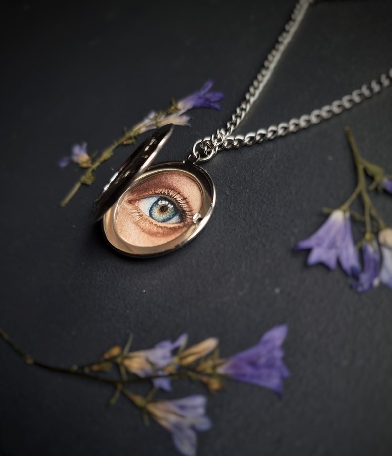 Op de afbeelding: Een zilveren ketting met een ovale hanger met een gedetailleerde schildering van een blauw en bruin oog. Het medaillon is open en onthult het kunstwerk. De ketting ligt op een donkere ondergrond, met takjes paarse bloemen verspreid.
