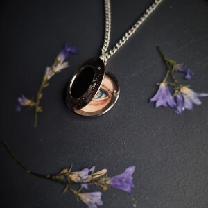 Op de afbeelding: Zilveren medaillonketting, geopend om een geschilderd oog met blauwe iris te onthullen. Het medaillon is omgeven door gedroogde paarse bloemen tegen een donkere achtergrond. Het medaillon heeft een decoratieve rand.