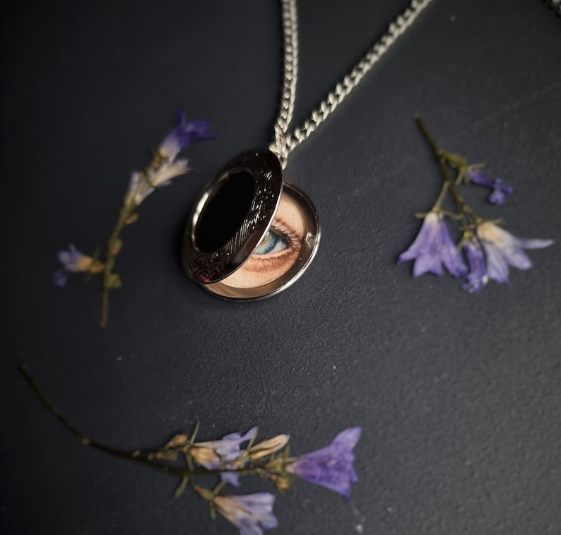 Op de afbeelding: Zilveren medaillonketting, geopend om een geschilderd oog met blauwe iris te onthullen. Het medaillon is omgeven door gedroogde paarse bloemen tegen een donkere achtergrond. Het medaillon heeft een decoratieve rand.