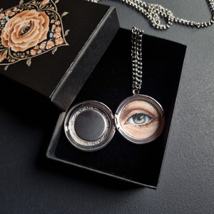 Op de afbeelding: Een zilveren ketting met een medaillon met een gedetailleerde schildering van een blauw oog, gepresenteerd in een open zwarte doos. De doosdeksel heeft een bloemenontwerp met een oog in het midden. De ketting is zilver.