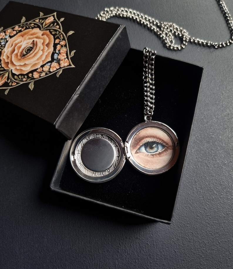 Op de afbeelding: Een zilveren ketting met een medaillon met een gedetailleerde schildering van een blauw oog, gepresenteerd in een open zwarte doos. De doosdeksel heeft een bloemenontwerp met een oog in het midden. De ketting is zilver.