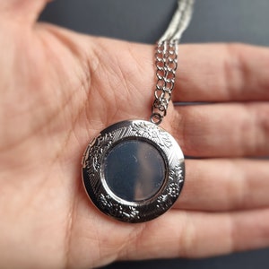 Op de afbeelding: Zilverkleurige ronde medaillonketting met een bloemmotief langs de randen. Het medaillon is open en toont een lege ruimte voor een foto. De ketting is ook zilverkleurig en delicaat.