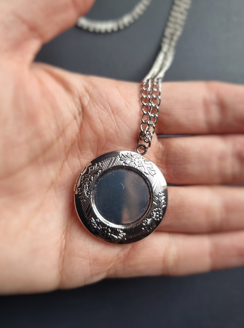 Op de afbeelding: Zilverkleurige ronde medaillonketting met een bloemmotief langs de randen. Het medaillon is open en toont een lege ruimte voor een foto. De ketting is ook zilverkleurig en delicaat.