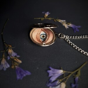 Op de afbeelding: Een zilveren ketting met een medaillon met een gedetailleerde oogillustratie. Het medaillon is open en toont de iris en huidtinten van het oog. De ketting is zilverkleurig en de achtergrond is donker met verspreide paarse bloemen.