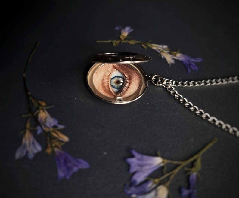 Op de afbeelding: Een zilveren ketting met een medaillon met een gedetailleerde oogillustratie. Het medaillon is open en toont de iris en huidtinten van het oog. De ketting is zilverkleurig en de achtergrond is donker met verspreide paarse bloemen.