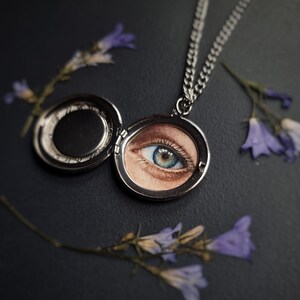 Op de afbeelding: Een zilveren medaillonketting met een open, cirkelvormige hanger met een gedetailleerde afbeelding van een blauw en bruin oog. De ketting ligt op een donkere ondergrond, met paarse bloesemtakjes rond de hanger.