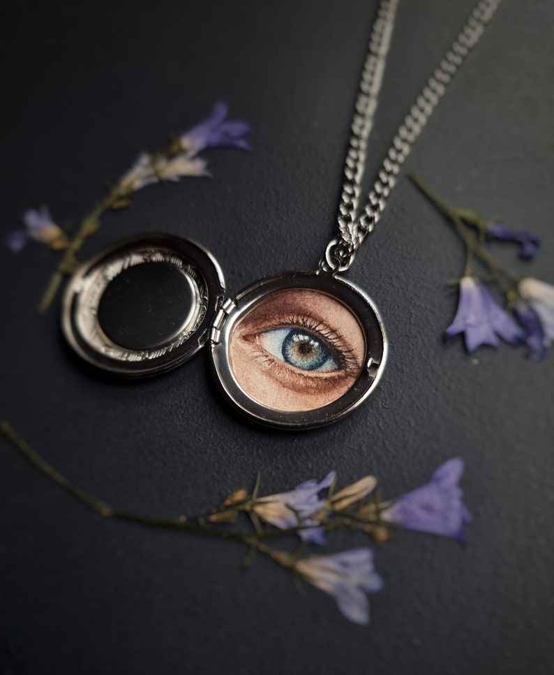 Op de afbeelding: Een zilveren medaillonketting met een open, cirkelvormige hanger met een gedetailleerde afbeelding van een blauw en bruin oog. De ketting ligt op een donkere ondergrond, met paarse bloesemtakjes rond de hanger.
