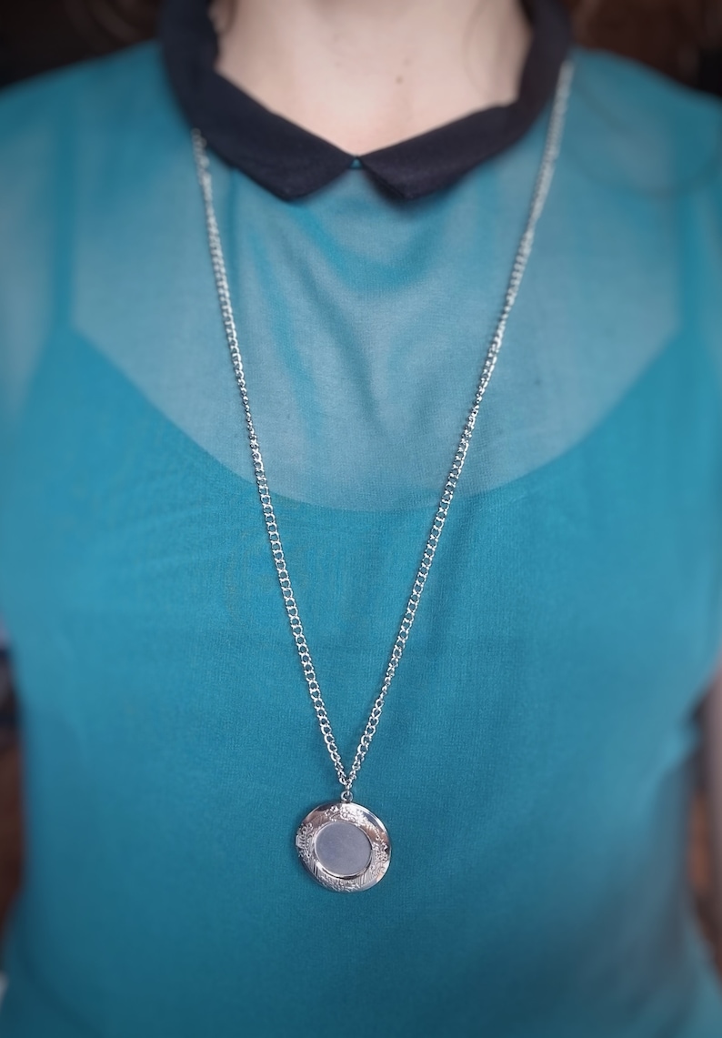 Op de afbeelding: Zilverkleurige ketting met een ronde hanger. De hanger heeft een decoratief bloemenpatroon langs de rand en een leeg midden. De ketting wordt gedragen over een turquoise top met een zwarte kraag.
