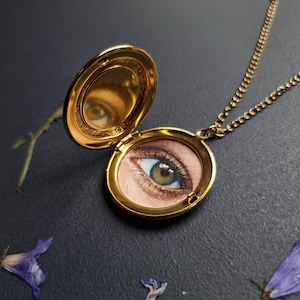 May include: A gold locket necklace with a painted eye inside. The eye is green with brown eyelashes and a pink eyelid.