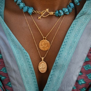 May include: A gold necklace with three strands and two pendants. The top strand is a turquoise stone necklace with a gold clasp. The middle strand has a round pendant with an intricate design. The bottom strand has an oval pendant with an intricate design.